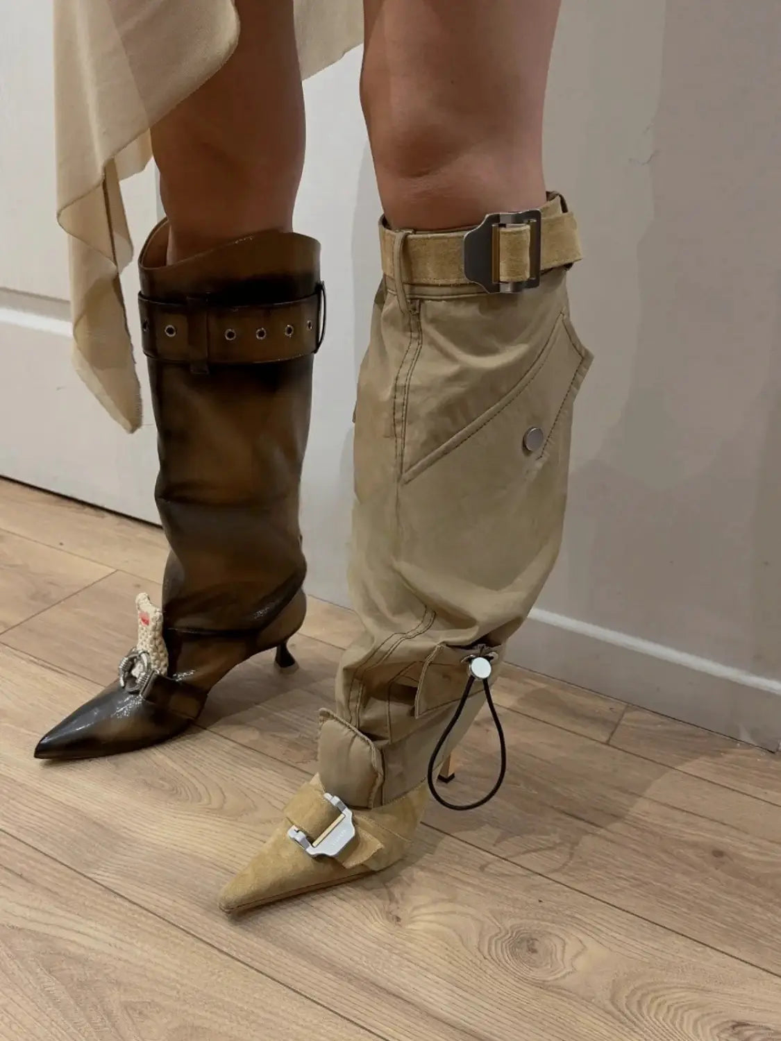 Two mismatched high heel pointed toe knee boots with buckles, one brown and one beige.