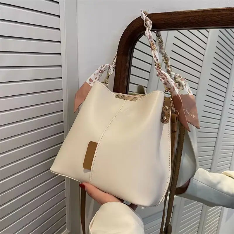 Cream-colored versatile bucket bag with brown leather trim and braided strap for women’s crossbody fashion.