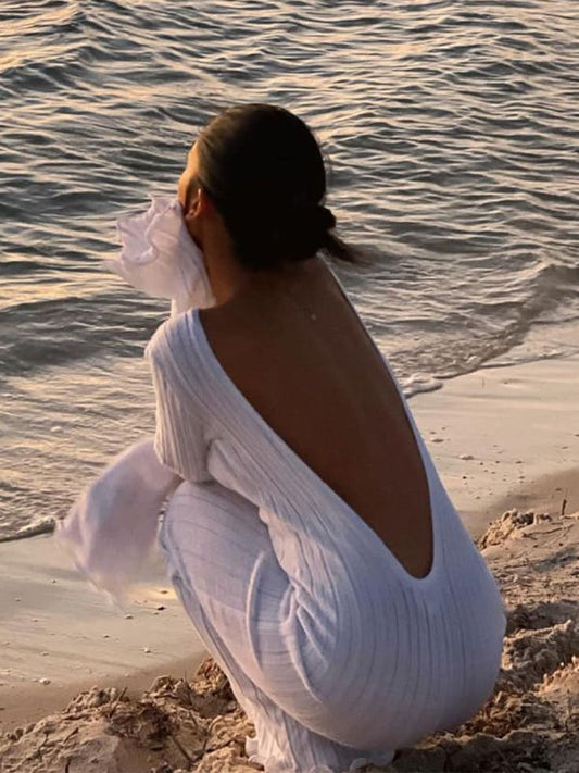 Person wearing a white dress sitting on a beach by the water.