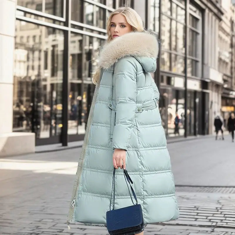 Long mint women’s puffer coat with luxurious fur-trimmed hood and navy handbag.