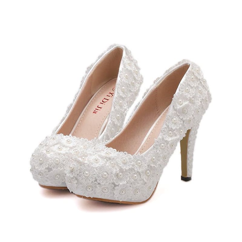 Pair of white lace high-heeled bridal shoes with floral embellishments.