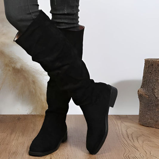 Pair of black knee-high boots with flat soles worn over skinny jeans.